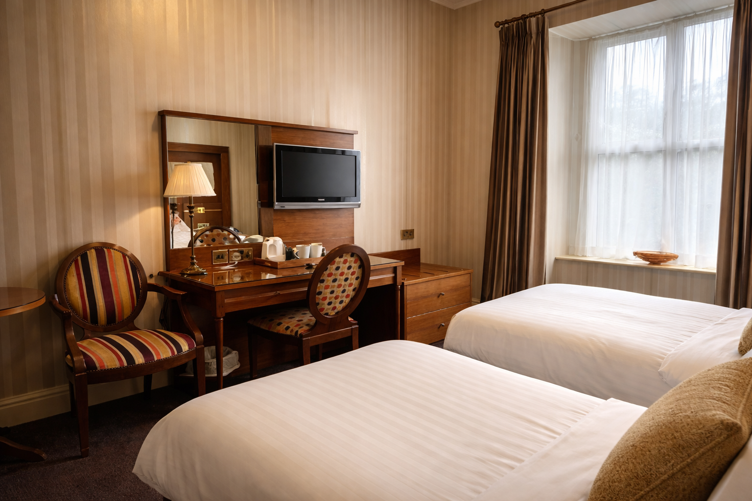 Twin hotel room with two single beds, wooden desk with mirror and TV, patterned chairs, and a large window with curtains.